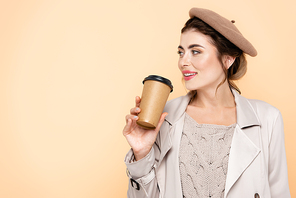 fashionable woman in autumn outfit holding coffee to go while looking away isolated on peach
