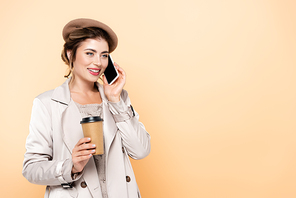 young woman in stylish autumn outfit holding coffee to go while talking on smartphone isolated on peach