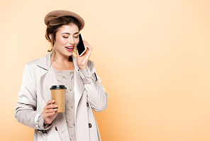 trendy woman in autumn outfit talking on mobile phone while holding coffee to go isolated on peach