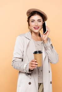 stylish woman in autumn outfit holding coffee to go while talking on smartphone isolated on peach