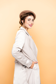 trendy woman in beret and trench coat  while posing with hand in pocket on peach