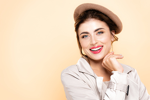 joyful, fashionable woman in beret holding hand near chin while   isolated on peach