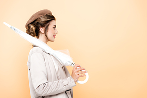 side view of fashionable woman in trench coat and beret holding folded umbrella on peach