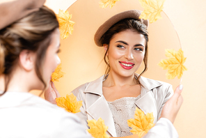 selective focus of young woman in trench coat and beret looking in mirror decorated with autumn leaves on peach