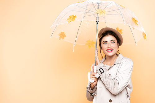 fashionable woman in trench coat and beret posing under umbrella with yellow leaves on peach