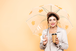 joyful woman in stylish autumn outfit holding coffee to go under umbrella with foliage on peach