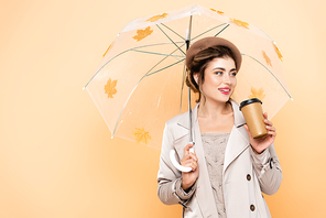 trendy woman in beret and trench coat holding coffee to go under umbrella with autumn leaves on peach
