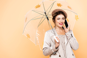 joyful woman in fashionable autumn outfit talking on mobile phone under umbrella decorated with yellow leaves on peach