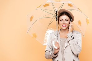 young woman in trendy autumn outfit talking on smartphone under umbrella with autumn leaves on peach