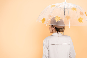 back view of trendy woman in beret and trench coat under umbrella, decorated with yellow leaves on peach