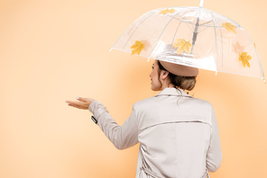 back view of fashionable woman standing with outstretched hand under umbrella, decorated with yellow leaves on peach