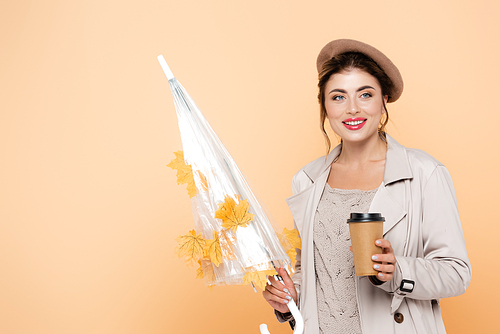 trendy woman in autumn outfit holding coffee to go and umbrella with yellow leaves on peach