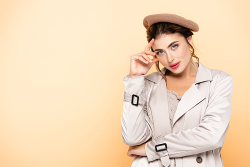 fashionable woman in trench coat and beret touching forehead while  on peach