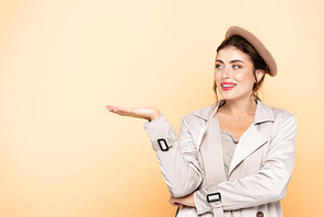 fashionable woman in trench coat and beret pointing with hand on peach