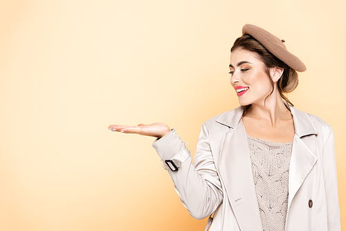 joyful woman in trench coat and beret pointing with hand on peach