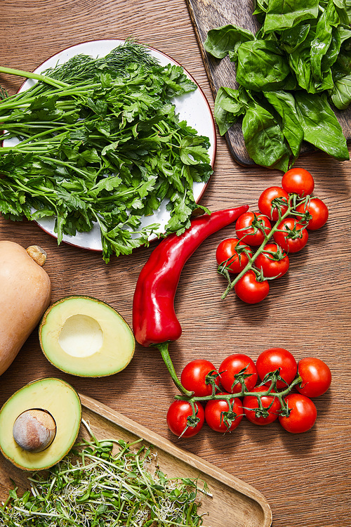 Top view of vegetables, avocado halves, greenery, microgreens and basil leaves on cutting boards on wooden background