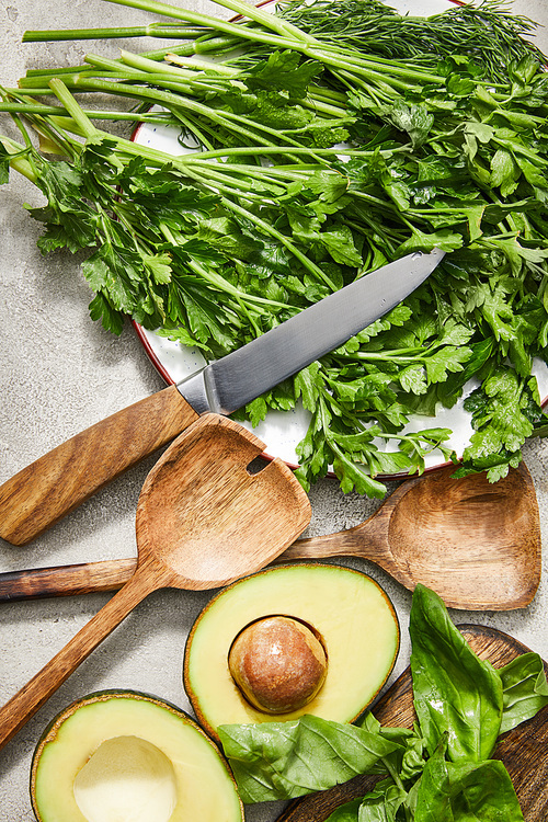 Top view of greenery on plate, knife, spatulas, avocado halves and basil leaves on grey background