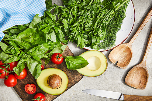 Top view of greenery, basil leaves, cherry tomatoes and avocado halves near spatulas, knife and cloth on grey background