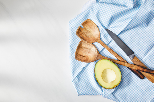 Top view of spatulas, knife and avocado half on plaid cloth on white background