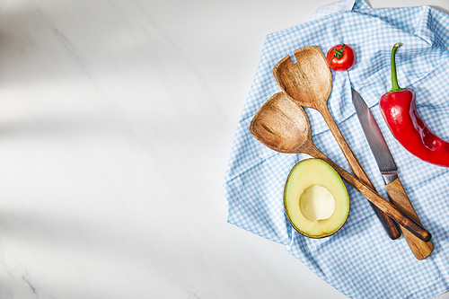 Top view of spatulas, knife, cherry tomato, chili pepper and avocado half on plaid cloth on white background