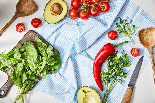 Top view of vegetables, greenery, avocado halves, knife and spatulas on plaid cloth near basil on cutting board on white