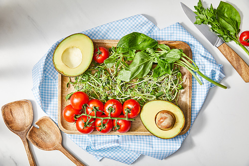 Top view of cutting board with microgreens, basil, cherry tomatoes and avocado on cloth near knife and spatulas on white