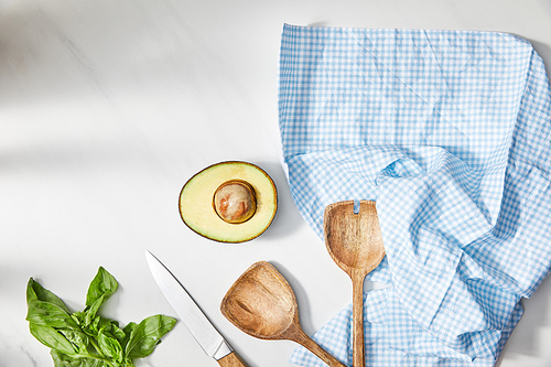 Top view of basil, knife, spatulas and avocado half near plaid cloth on white background