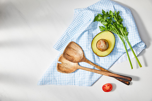 Top view of parsley and spatulas near avocado and cherry tomato halves with plaid cloth on white background