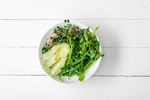top view of arugula, avocado and microgreen in bowl on white wooden surface