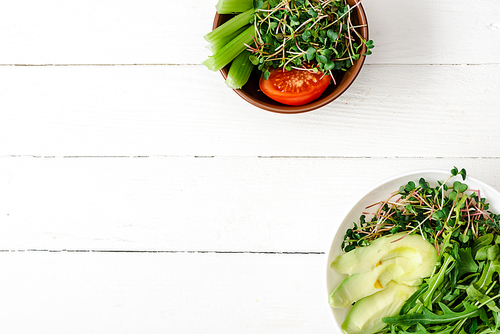 top view of fresh vegetables with avocado and microgreen in bowls on white wooden surface