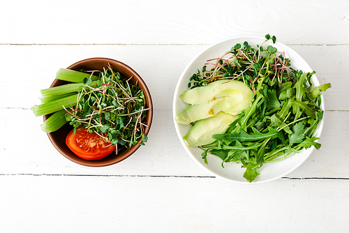 top view of fresh vegetables with avocado and microgreen in bowls on white wooden surface