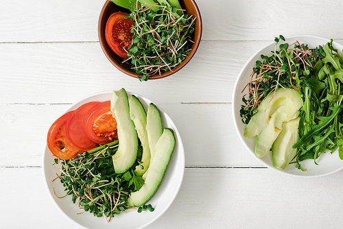 top view of fresh vegetables with avocado and microgreen in bowls on white wooden surface