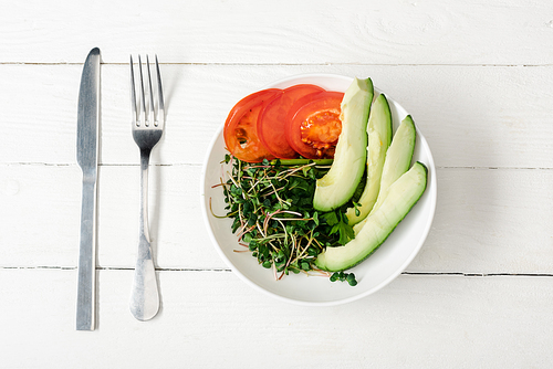 top view of tomato, avocado and microgreen in bowl on white wooden surface with cutlery