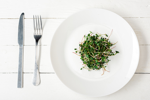 top view of fresh microgreen on plate near cutlery on white wooden surface