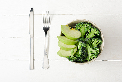 top view of fresh green apple and broccoli in bowl near cutlery on white wooden surface