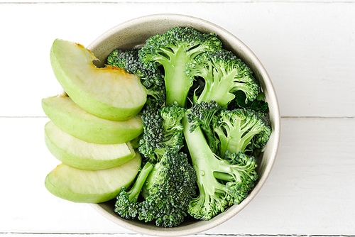 top view of fresh green apple and broccoli in bowl on white wooden surface