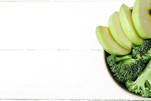 top view of fresh green apple and broccoli in bowl on white wooden surface