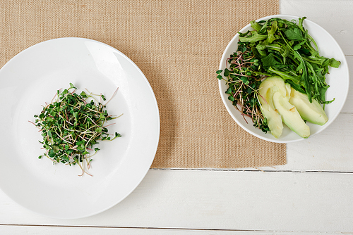 top view of fresh microgreen on plate near bowl of green salad on beige napkin on white wooden surface