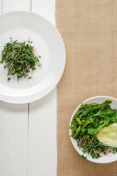 top view of fresh microgreen on plate near bowl of green salad on beige napkin on white wooden surface
