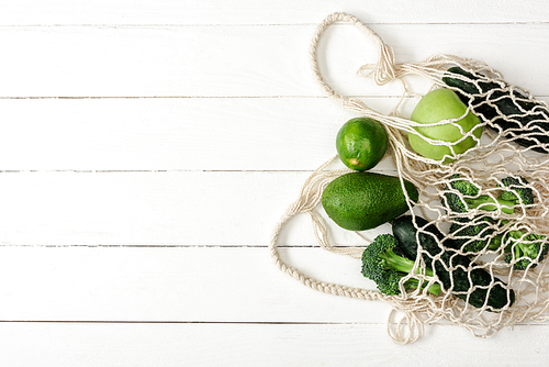 top view of fresh green fruits and vegetables in string bag on white wooden surface