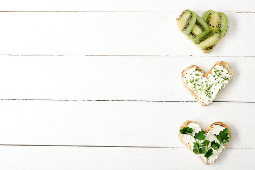 top view of heart shaped canape with creamy cheese, microgreen, parsley and kiwi on white wooden surface