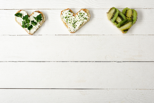 top view of heart shaped canape with creamy cheese, microgreen, parsley and kiwi on white wooden surface