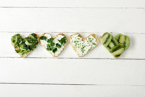 top view of heart shaped canape with creamy cheese, broccoli, microgreen, parsley and kiwi on white wooden surface