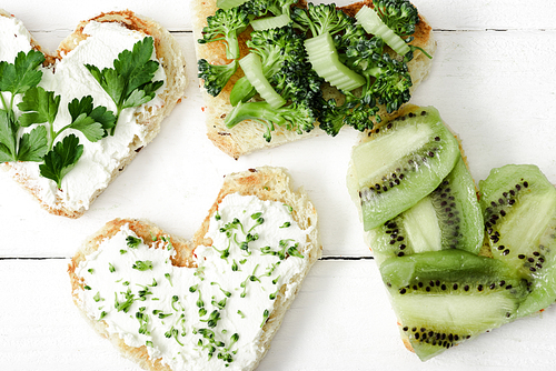 top view of heart shaped canape with creamy cheese, broccoli, microgreen, parsley and kiwi on white wooden surface