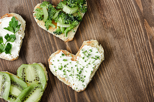 top view of heart shaped canape with creamy cheese, broccoli, microgreen, parsley and kiwi on wooden surface