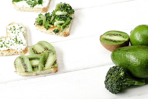 heart shaped canape with creamy cheese, broccoli, microgreen, parsley and kiwi near green fruits and vegetables on white wooden surface
