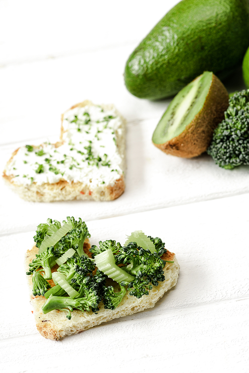 selective focus of heart shaped canape with creamy cheese, broccoli, microgreen near green fruits and vegetables on white wooden surface