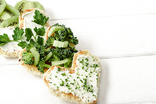 heart shaped canape with creamy cheese, broccoli, microgreen, parsley and kiwi on white wooden surface