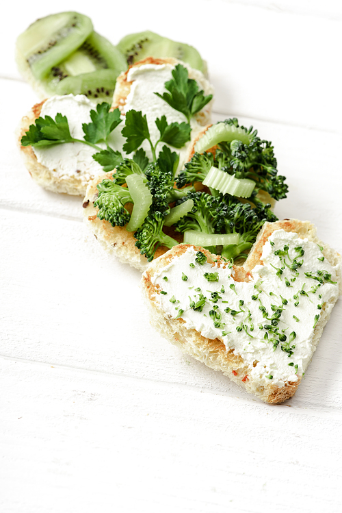 selective focus of heart shaped canape with creamy cheese, broccoli, microgreen, parsley and kiwi on white wooden surface