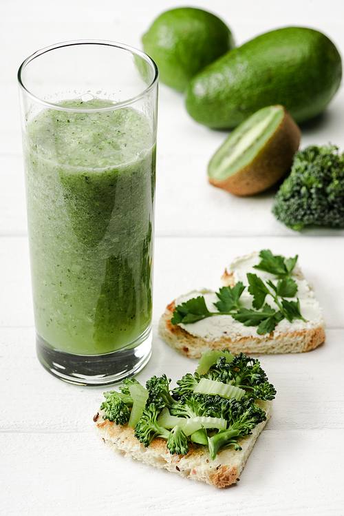 selective focus of green smoothie and heart shaped canape with creamy cheese, broccoli, parsley near fruits and vegetables on white wooden surface